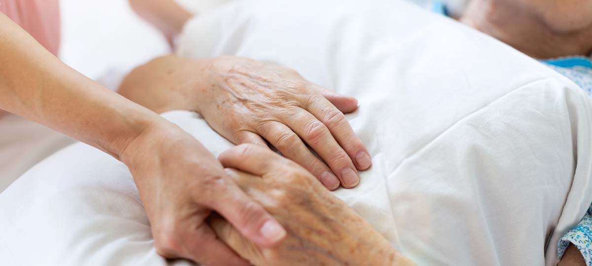 Young hand holding older hands of patient laying down in hospital bed