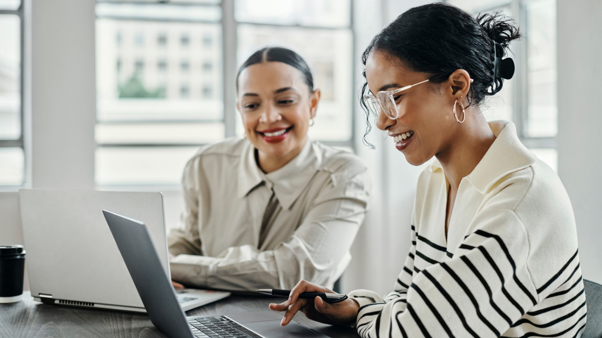 An image of two women sitting in an office, laptops on the table in front of them. Both women are smiling at their screens.