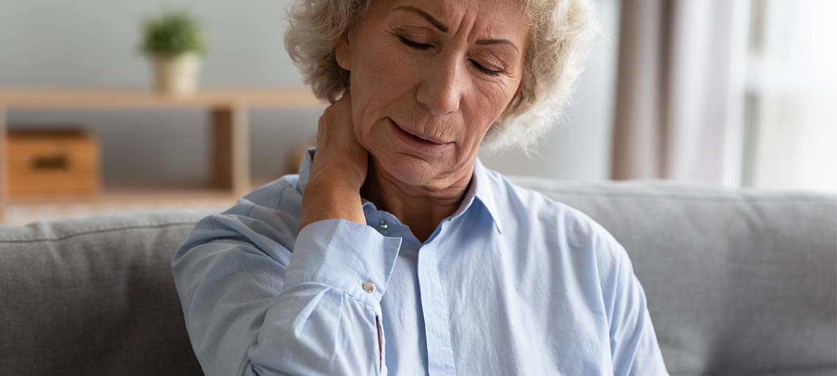 Older woman sitting on couch with painful expression and hand on neck